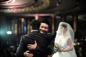 Groom sharing an emotional hug with a guest while bride in white gown stands in background.