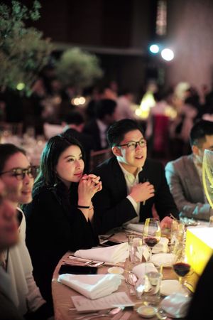 Elegantly dressed wedding guests seated at a candlelit reception dinner table in a grand ballroom.