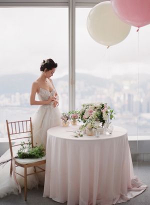 Bride in strapless gown stands by floor-to-ceiling windows with pink balloons and floral centerpiece.