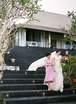 Bride in lace gown descends black stone stairs with woman in lavender dress at tropical venue.