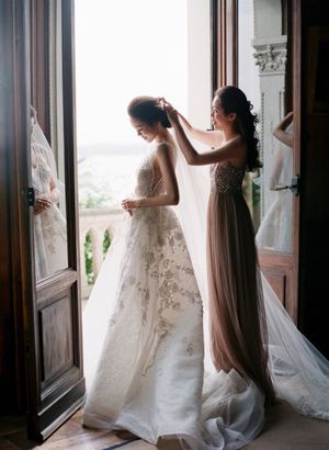 Bridesmaid in taupe gown adjusts bride's veil in sunlit doorway with ocean view backdrop.