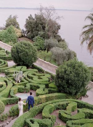 Aerial view of bride and groom walking through ornate hedge maze garden beside a lake.