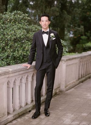 Groom in black tuxedo with boutonniere poses confidently on stone balustrade in garden setting.