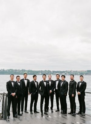 Groom and groomsmen in black tuxedos pose together on waterfront dock under overcast skies.