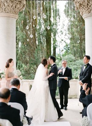 Bride and groom share first kiss at outdoor ceremony beneath hanging crystal globe decorations.
