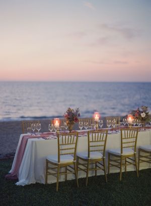 Romantic beachside wedding reception table set with candles and florals at sunset.