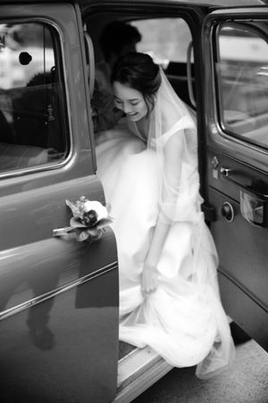 Black and white photo of a smiling bride in veil stepping out of a vintage car.