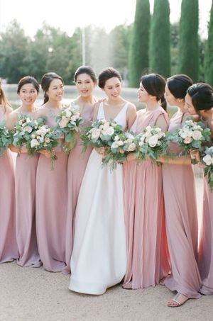 Bride in white gown surrounded by bridesmaids in dusty rose dresses holding white bouquets.