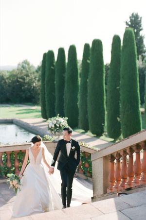 Bride and groom hold hands walking up stone steps at elegant garden wedding venue with tall cypress trees.
