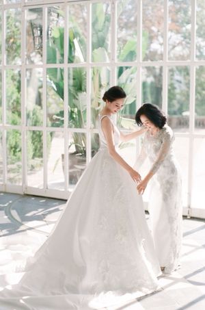 Bride in floral ballgown holds hands with mother in sunlit greenhouse during wedding day preparations.
