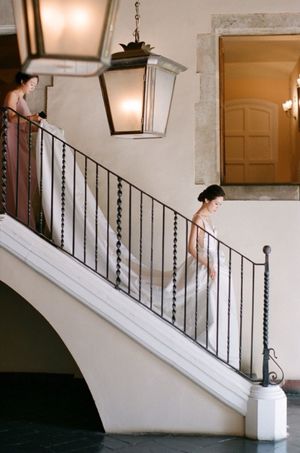 Elegant bride descends grand staircase with flowing veil in classic villa interior with wrought iron railing.