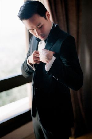 Dapper groom in black tuxedo adjusting his cufflinks by a window before the wedding.