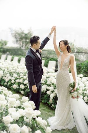 Groom twirls bride in beaded mermaid gown amid white floral aisle at outdoor garden wedding ceremony.