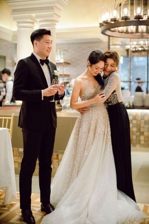 Bride in sparkling ballgown shares emotional embrace with guest while groom holds champagne at reception.