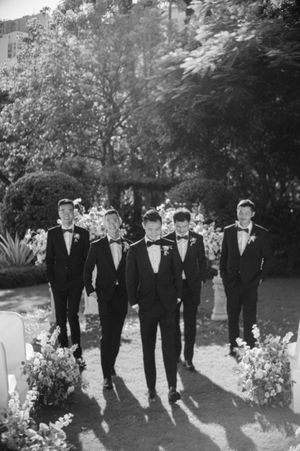 Black and white photo of groom and groomsmen in black tuxedos walking through garden wedding venue.