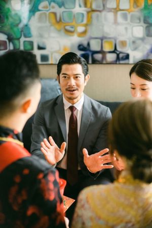 Groom in gray suit gestures expressively during traditional Chinese wedding tea ceremony with family.