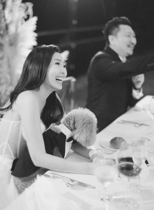 Bride laughing joyfully at wedding reception table holding a small dog dressed in a tuxedo.