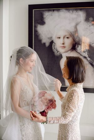 Bride in lace wedding gown and veil receiving bouquet of pink roses from a woman indoors.