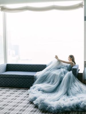 Bride in a voluminous blue tulle ball gown sitting on a sofa by a large window with city views.