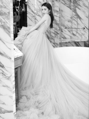 Elegant bride in a flowing tulle gown with ruffled details standing by a marble bathtub in black and white.
