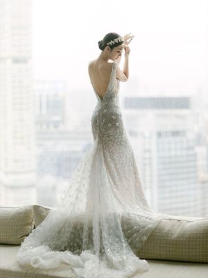 Bride in a backless floral lace wedding gown with train standing by a panoramic city window.