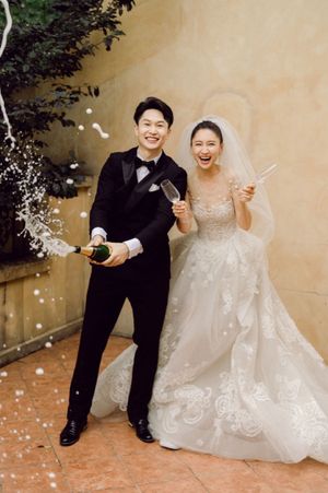 Happy couple popping champagne in wedding attire, bride in lace gown with veil, groom in black tuxedo.