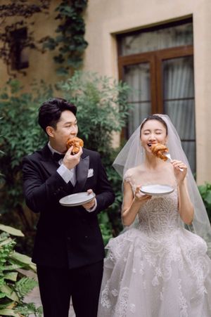 Bride and groom in wedding attire enjoying food outdoors in a lush garden setting.