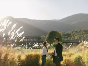 Couple holding hands in a golden meadow with mountains and sunset in the background.