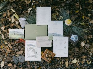 Wedding stationery flat lay with sage green invitation suite on pebbles with pine accents.