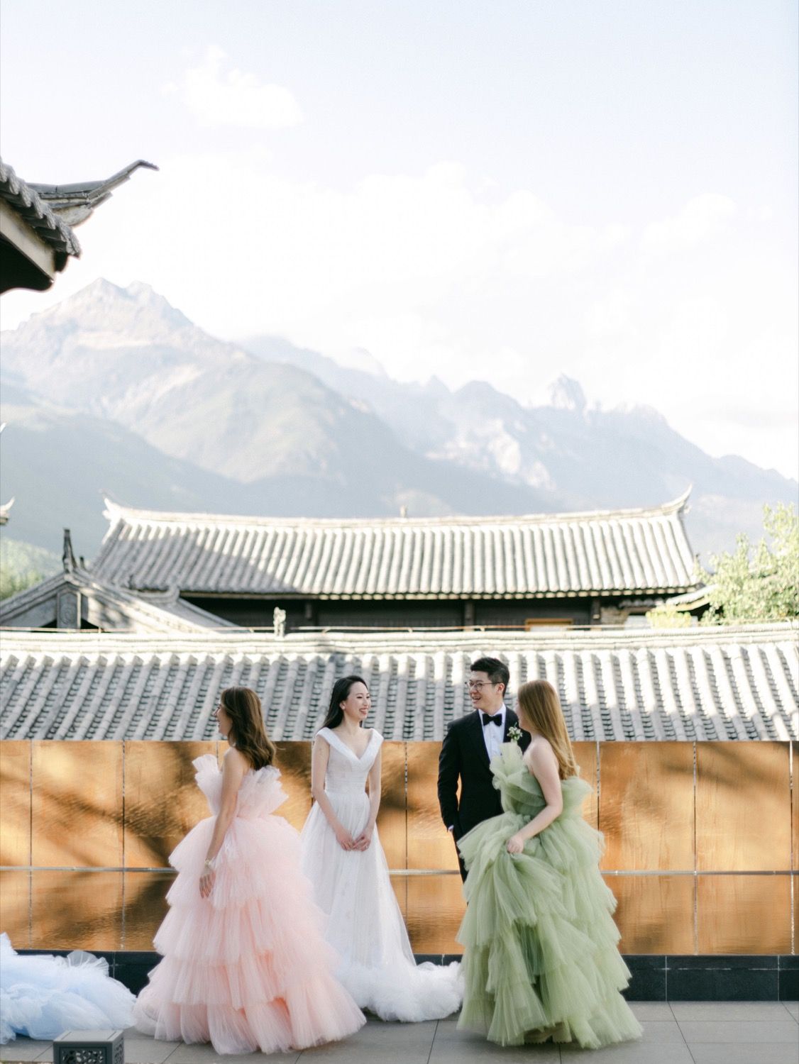 Bride and wedding party in colorful gowns pose before traditional Chinese rooftops with mountain backdrop.