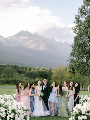 Bride, groom, and bridesmaids in mismatched pastel gowns pose outdoors with stunning mountain scenery.