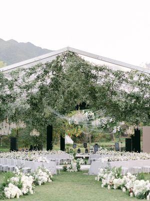 Elegant outdoor wedding reception under a glass tent adorned with lush greenery and white floral arrangements.