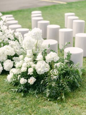 White roses and hydrangeas with lush greenery line the aisle beside cylindrical white ceremony seats.