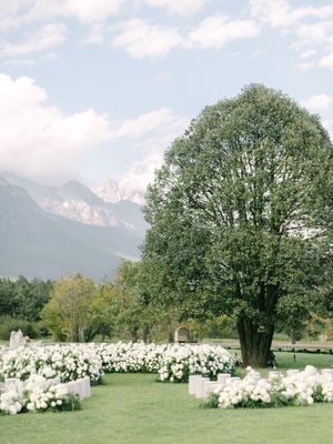 Outdoor wedding ceremony setup with white floral rows and a grand tree against mountain backdrop.