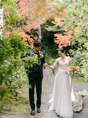 Bride and groom walk hand in hand on a garden path surrounded by vibrant autumn foliage.