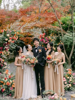 Groom stands smiling with four bridesmaids in gold dresses amid colorful floral arrangements.