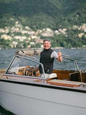 Smiling man giving thumbs up while steering a classic wooden speedboat on a scenic lake.