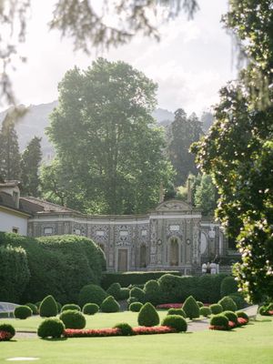 Ornate European villa garden with manicured topiary hedges and red flowers framed by lush trees.