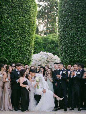 Groom dips bride for a romantic kiss as wedding party celebrates between tall green hedgerows.