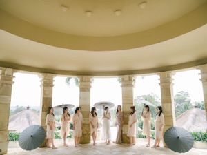 Bridesmaids in blush dresses hold black parasols under a columned pavilion overlooking lush scenery.