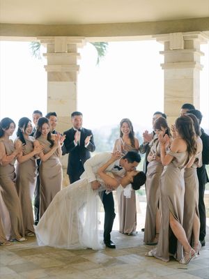 Groom dips bride for a kiss surrounded by cheering wedding party in a sunlit colonnade.