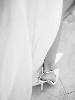 Black and white close-up of bride's crystal-adorned heel peeking beneath a flowing white gown.