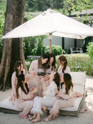 Bridesmaids in blush robes celebrating with champagne on a beach daybed under a white umbrella.