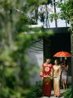 Couple in traditional Chinese wedding attire walking through a tropical garden with a red umbrella.