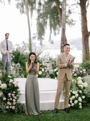 Wedding emcees in elegant attire speaking at an outdoor ceremony adorned with white floral arrangements.