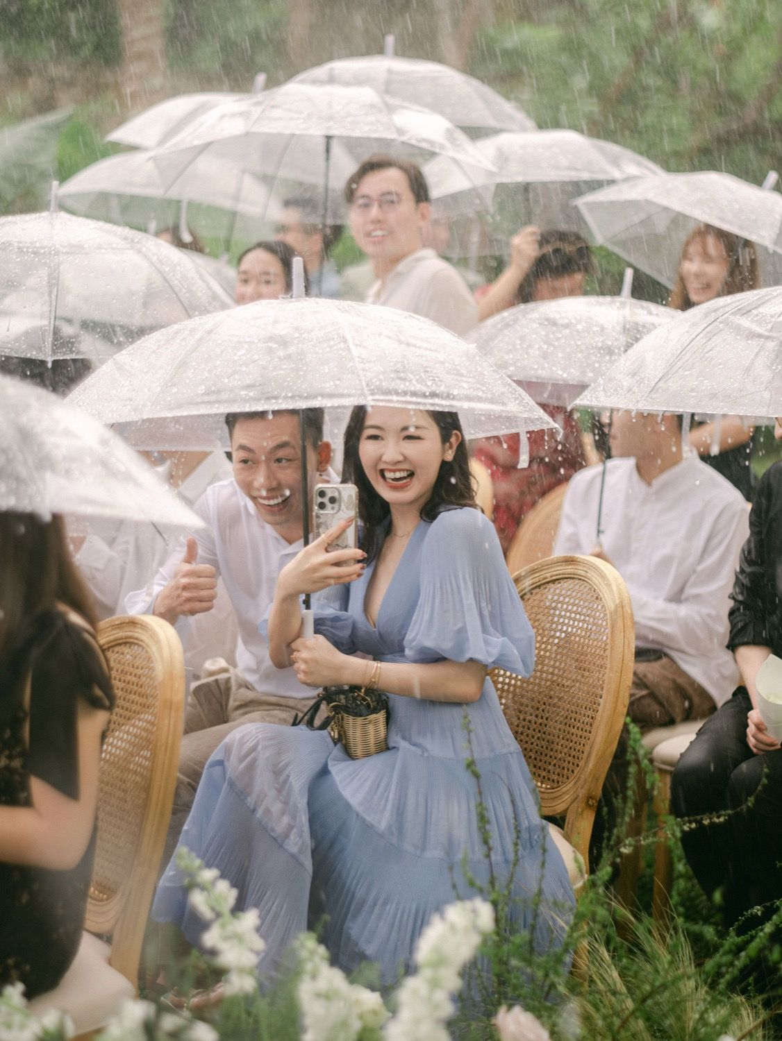 Wedding guests laughing under clear umbrellas in the rain at an outdoor garden ceremony.