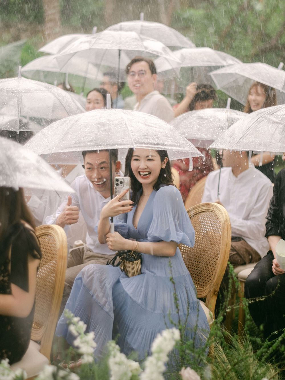 Wedding guests laughing under clear umbrellas in the rain at an outdoor garden ceremony.