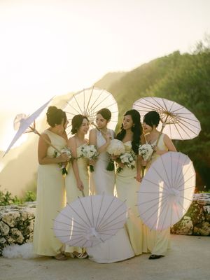 Bridesmaids in yellow dresses holding white parasols pose with bride at golden hour outdoor wedding.