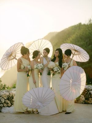 Bride and bridesmaids in yellow dresses laughing together holding white parasols at golden hour.