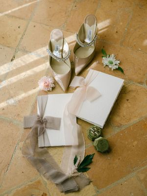 Elegant wedding details with Jimmy Choo heels, ribbon-tied vow books, and floral accents on tile.
